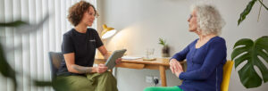 female patient sat at a table in consultation with a practitioner who holds a tablet
